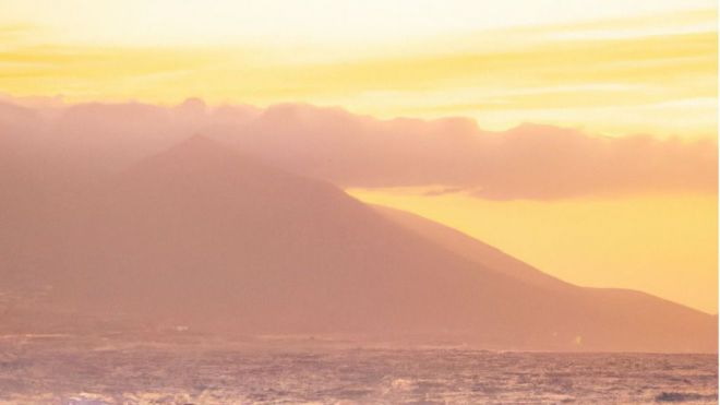 En una playa de Canarias, una pareja camina por la arena blanca mientras el sol se pone en el horizonte. El cielo está teñido de tonos naranjas y rosados, creando un paisaje espectacular. A lo lejos, se pueden ver las montañas volcánicas que caracterizan a las islas Canarias. La pareja se detiene para admirar el paisaje y aprovecha para tomar fotografías de este momento mágico. A pesar del clima cálido y seco, la brisa marina refresca el ambiente y crea una sensación de tranquilidad.